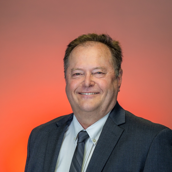 Kenneth Menkhaus smiles in a professional headshot, wearing a dark gray suit jacket, a light dress shirt, and a gray patterned tie against an orange and neutral gradient background.