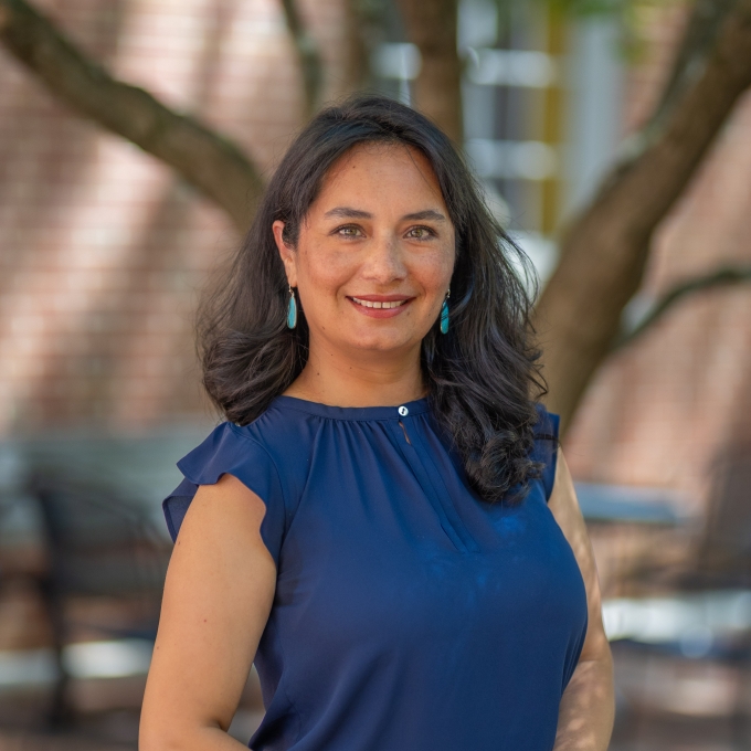 Portrait of Marcela Bacon wearing a dark blue short-sleeved top and turquoise drop earrings, smiling outdoors.
