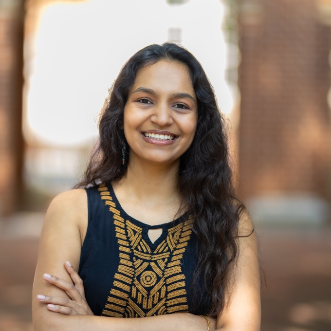 A smiling young woman with long, dark, wavy hair and arms crossed in a sunlit outdoor courtyard