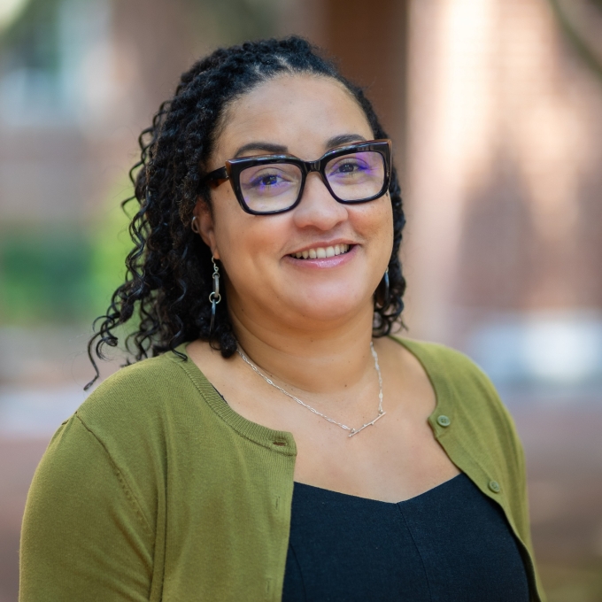 Trasheil Sinnett smiles in a headshot, wearing thick black-framed glasses, dark braided hair, and an olive green cardigan over a black top, with a softly blurred background.