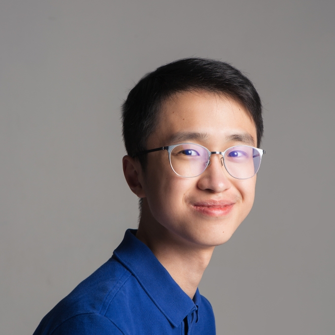 A male student with glasses and a blue button down smiles softly at the camera with head turned