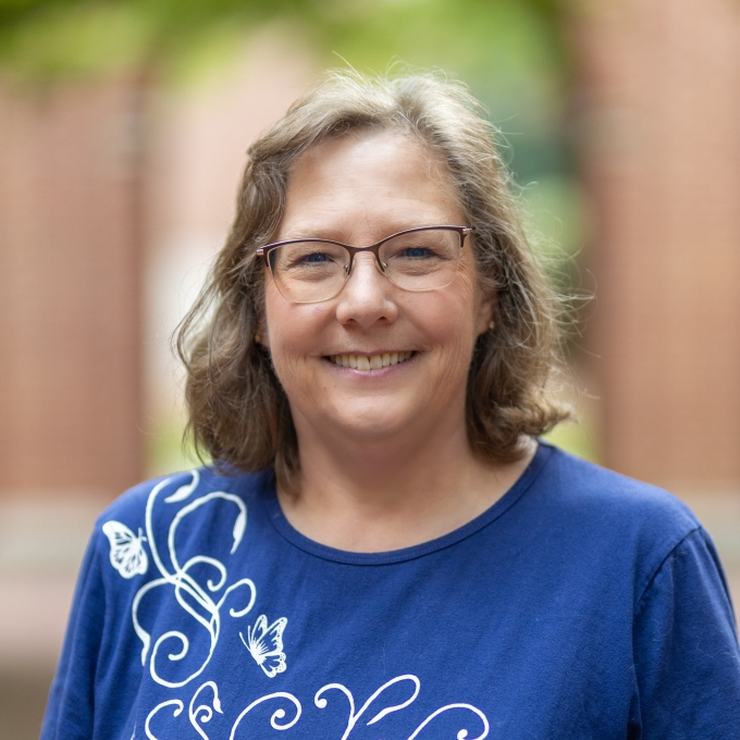 A smiling woman with light brown hair, glasses, a blue embroidered blouse stands in an outdoor courtyard