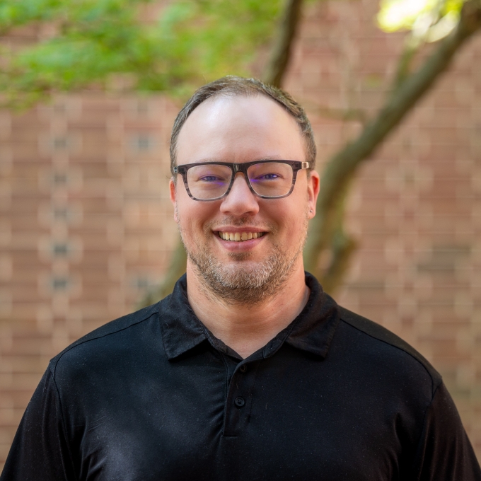A man with glasses, short light facial hair, and a black polo smiles in an outdoor courtyard