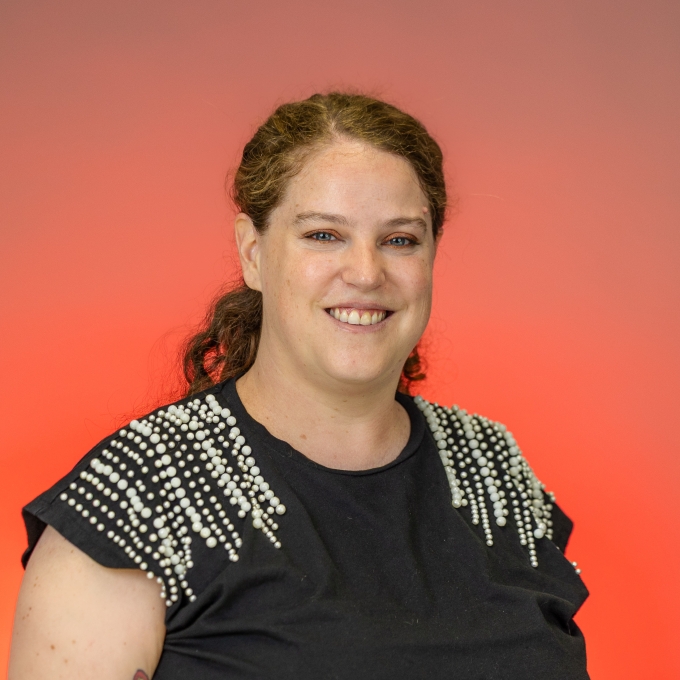 A person with a dark, beaded shirt, red curly hair, hazel eyes and light skin on a red background