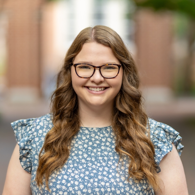 A smiling young woman with long, wavy brown hair and brown tortise-shell glasses in an outdoor courtyard