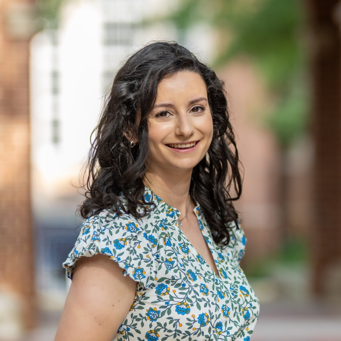 A woman with shoulder-length, dark curly hair and a floral blouse smiles brightly against a blurred outdoor courtyard