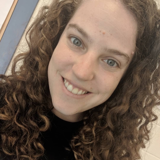 A person with long, curly brown hair and hazel eyes smiling directly at the camera.