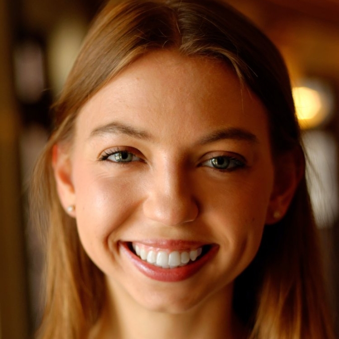 Close-up portrait of a smiling young woman with light brown hair, wearing a pattern-printed top.