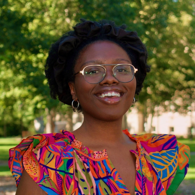 A Black woman wearing wire-rimmed glasses, silver hoop earrings, and a brightly colored tropical-print top smiles outdoors with lush green trees in the background.