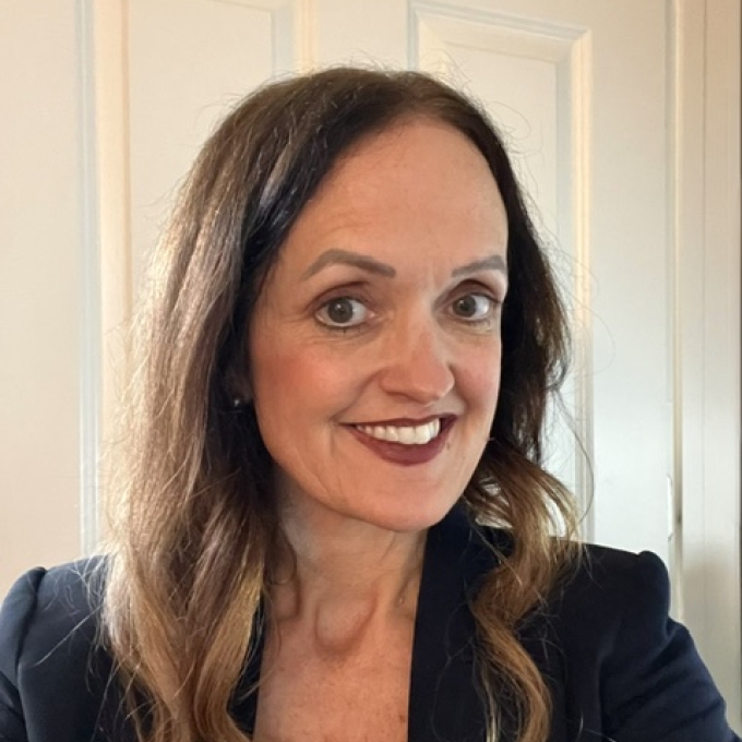 A woman with long, wavy brown hair with light ends smiles while wearing a dark blazer over a green top in front of a white door.