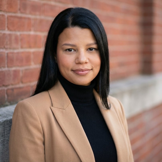 A smiling woman with shoulder-length dark hair wearing a camel blazer and a black turtleneck standing in front of a red brick wall