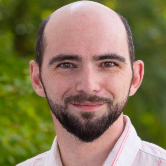 A smiling, light-skinned man with a bald head, dark brown eyes, and a full dark beard against a blurred green background.
