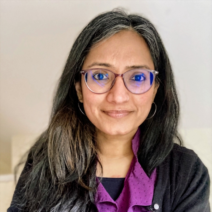 A headshot of a woman with long grey-streaked dark hair, wearing glasses, thin hoop earrings, and a purple patterned top paired with a black cardigan.