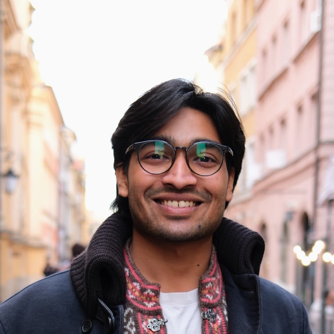 A male student wtih a navy blue jacket and colorful shirt, glasses, standing on a city street