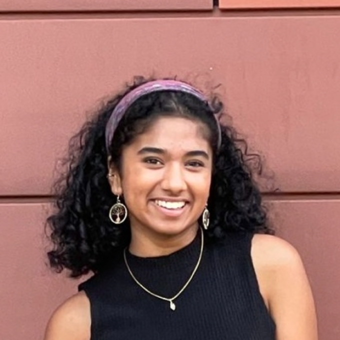 A woman with dark curly, shoulder-length hair in a purple headband stands in front of a red terra cotta wall