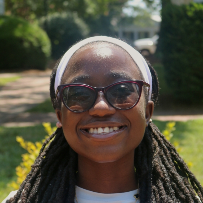 A smiling female student with glasses, white shirt, long dark hair in intricate braids and headband