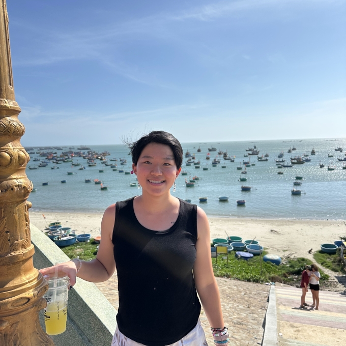 A smiling person with short dark hair and a black tank top holds a cold drink while standing on stairs overlooking a sandy beach dotted with round coracle boats and floating vessels on clear blue ocean water.