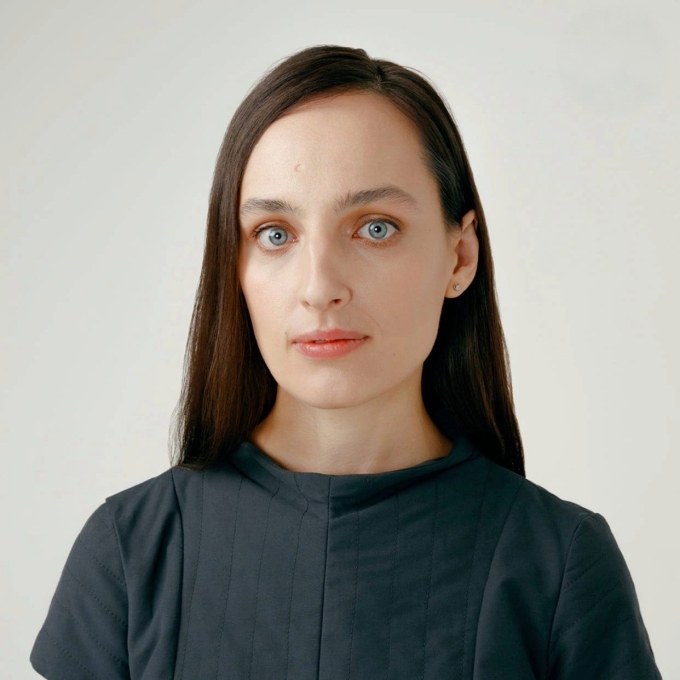 A woman with long dark hair and light eyes looking directly forward, wearing a dark short-sleeved top against a plain light background
