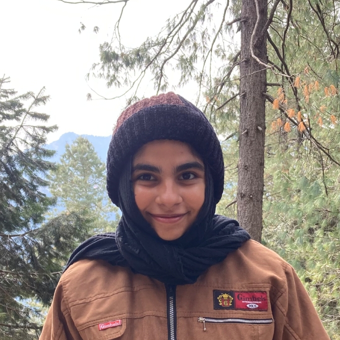 A female student in a forest with a hat and scarf covering her hair, brown canvas jacket, and a mountain view in the background