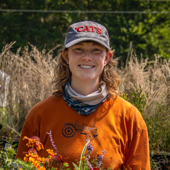 A woman with a 'Cats baseball cap, orange t-shirt, neck gaitor, and shoulder-length blonde curly hair smiles in a greenhouse