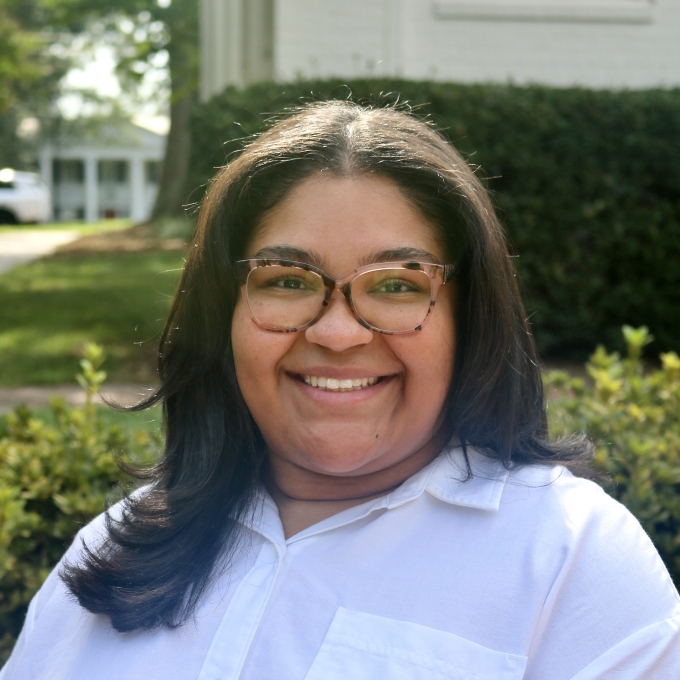 Mariah smiling outdoors wearing glasses and a white collared shirt with shoulder-length dark hair.