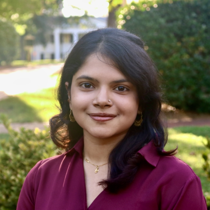 Mishti smiling outdoors in a deep plum-colored collared shirt with shoulder-length dark hair