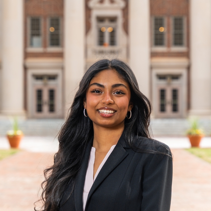 A smiling woman with long dark wavy hair wearing a black blazer over a light shirt standing in front of a brick building.