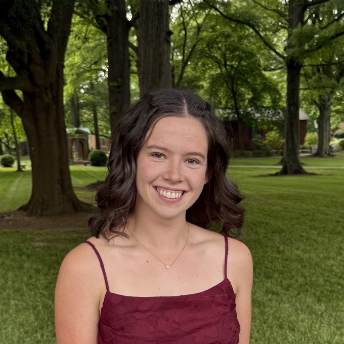 A smiling woman with dark, shoulder-length wavy hair wears a maroon dress and necklace while posing outdoors in front of tall trees