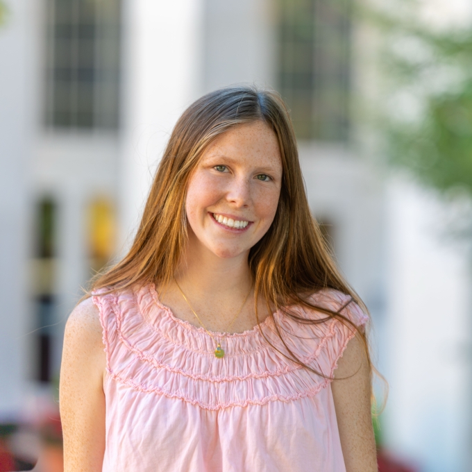 A smiling woman with long straight light brown hair wearing a sleeveless pink top and a gold pendant necklace outdoors