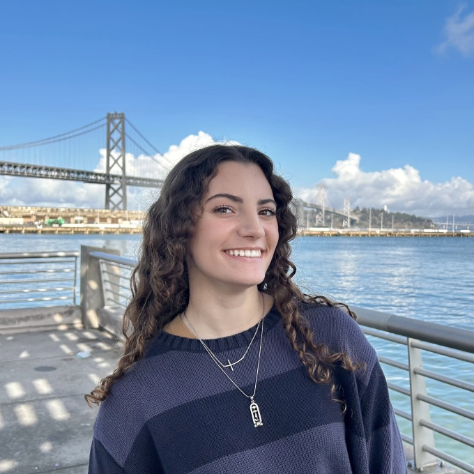 A smiling woman with long curly brown hair wears a dark striped sweater and layered necklaces while posing in front of a bay and bridge under a bright blue sky