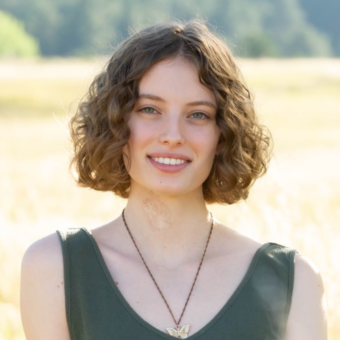 A woman with short, wavy light-brown hair and a butterfly necklace smiles in front of a sunlit grassy field