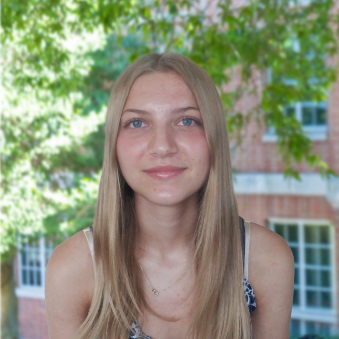 A smiling woman with long, straight blonde hair and light blue eyes wears a simple necklace in front of soft green foliage and a brick building