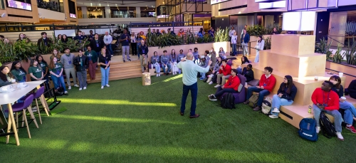 Students listen intently and sit in a modern atrium with a golf putting green carpet