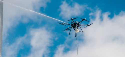 A high-altitude shot of a black multi-rotor drone tethered to the ground while emitting a powerful horizontal pressurized spray against a backdrop of blue sky and white clouds.