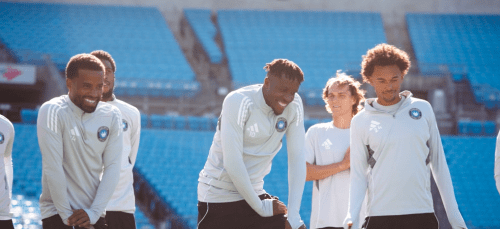 Five Charlotte FC soccer players wearing light grey training tops smile and warm up on a pitch in front of empty blue stadium seats.
