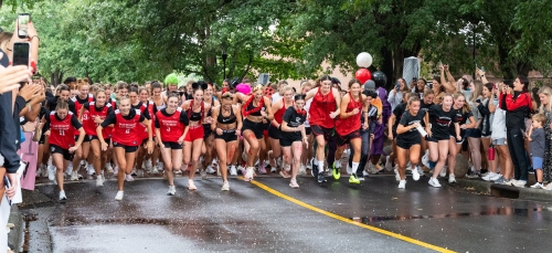 A large crowd of female college athletes in red and black athletic gear sprint down a wet, tree-lined street as spectators cheer and film from the sidewalks.