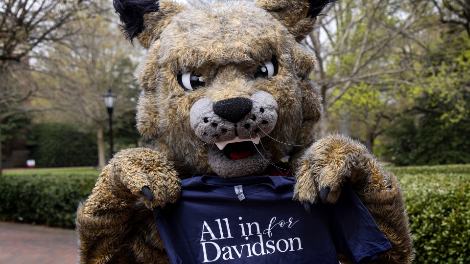 Mascot Lux the wildcat holds t-shirt that says "All in for Davidson" outside on campus