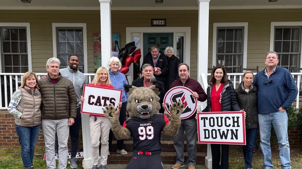 Peter Andrews, group of Davidson staff including president & athletic director, and Lux the wildcat outside Dave & Barbara Fagg's house with Davidson signs "Cats" and "Touch Down" in hands