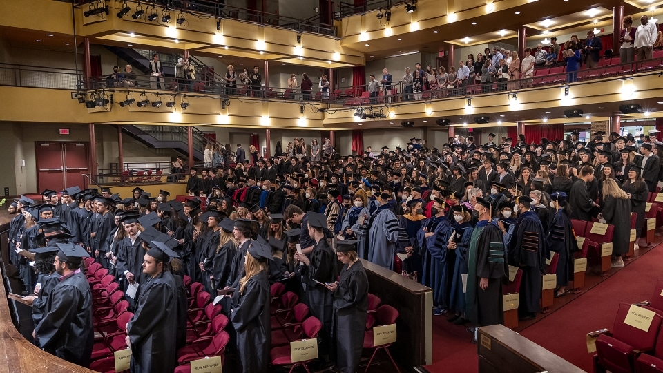 Students standing in rows of seats in cap & gowns in Duke Family Performance Hall