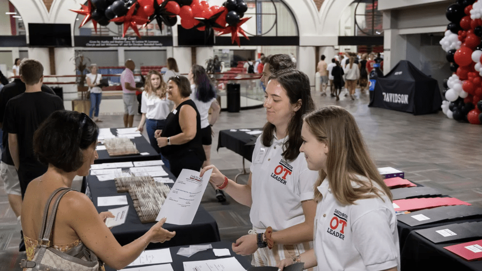 two young women in white polos talk to an older woman around a table with balloons in the background