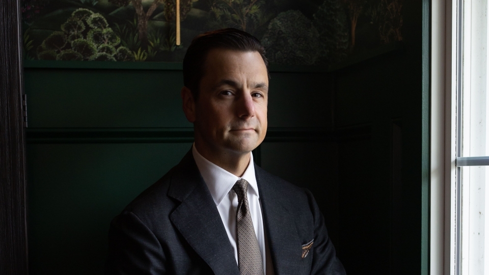 a young white man wearing a suit sitting in a dark room with a lamp behind