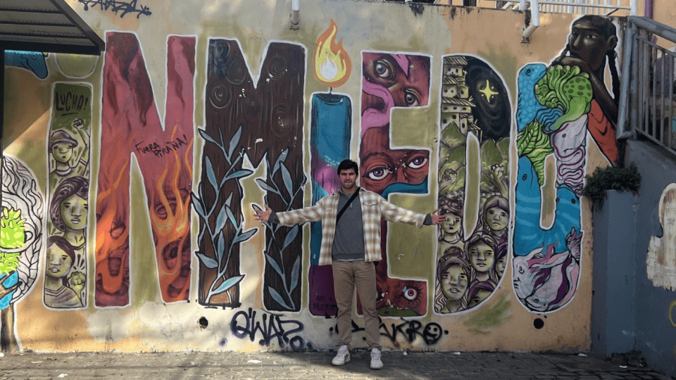 a young white man stands in front of a wall that reads " sin miedo"