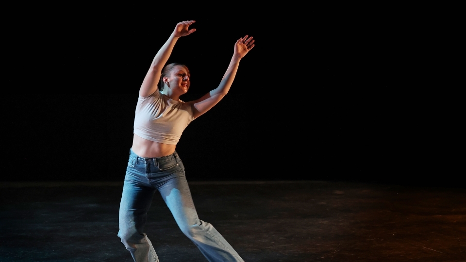 a young woman dances in a white top and jeans on a black stage