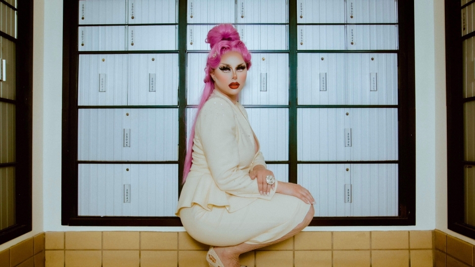 a young white woman in drag crouches in front of a wall of post office boxes