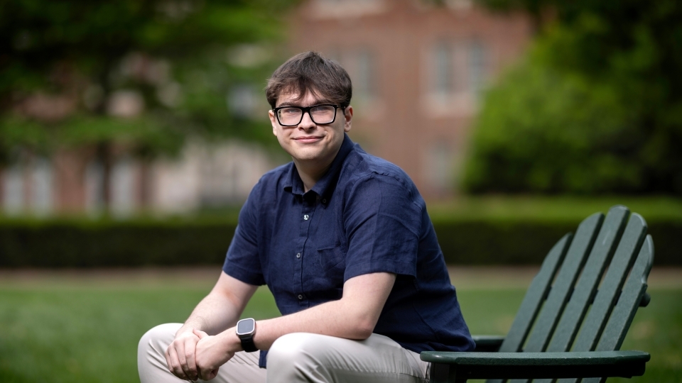 a young white man with a collared blue shirt sitting on a green adirondack chair on a college campus