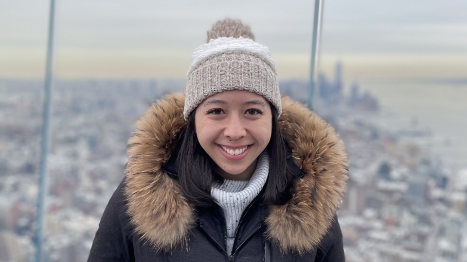 a young woman stands on the roof of a skyscraper in New York City, she wears a beanie and big coat and the sky appears to be grey and cloudy