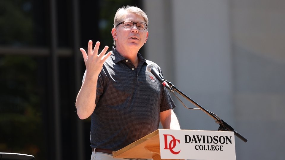 an older white man speaks at a lectern with "Davidson College" on it on a sunny day outside