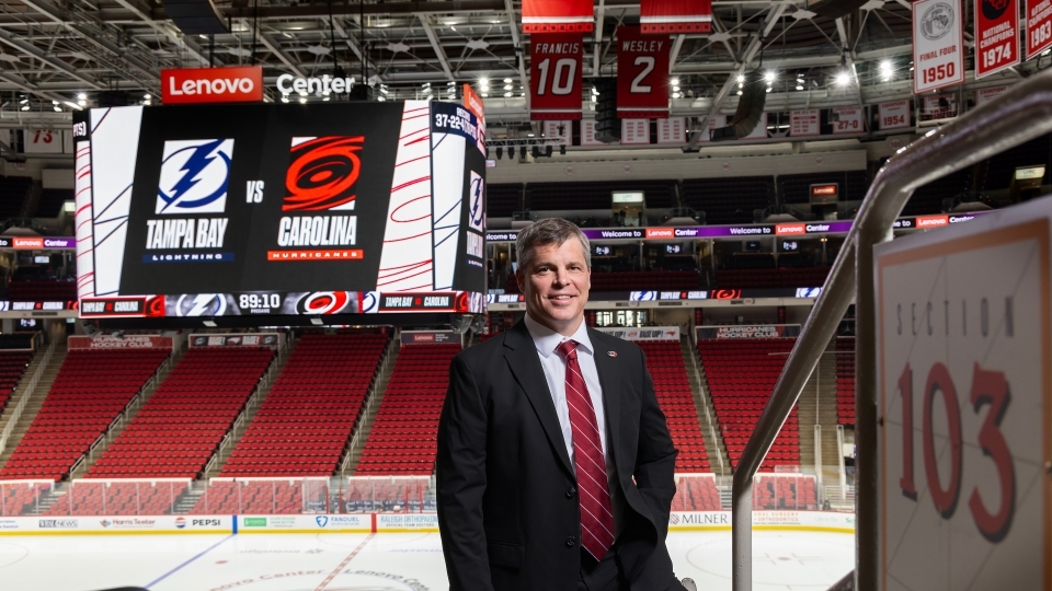 a middle aged white man wearing a black suit with a red tie in front of the Carolina Hurricanes hockey rink