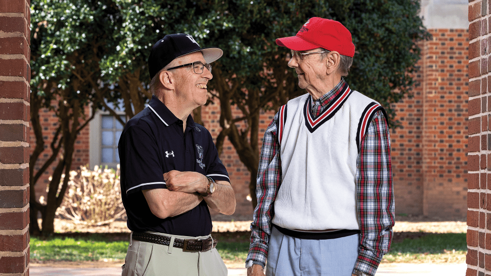 two older men stand together smiling looking at each other in front of a stadium, wearing Davidson College gear