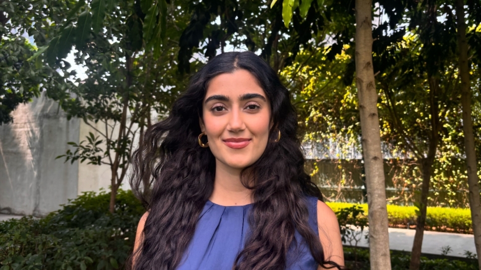 a young woman with long dark curly hair wearing a blue top standing outside in front of a set of green trees